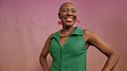 Portrait of woman with hands on hips against peach background. She is wearing a green top. 