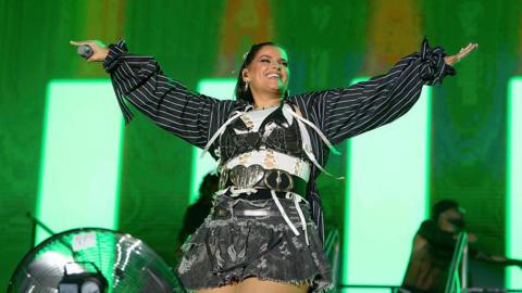 Nelly Furtado performs with a green light behind her and she is holding a microphone in her right hand and holding both arms up high.