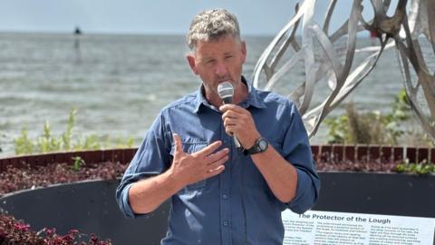 A grey haired man wearing a blue shirt is speaking into a microphone on the shore of Lough Neagh