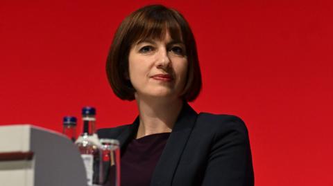 Education secretary Bridget Phillipson, who has short brown hair and a fringe, wears a black blazer and dark top and is pictured against a red background. Bottles and a glass of water are seen in the foreground.