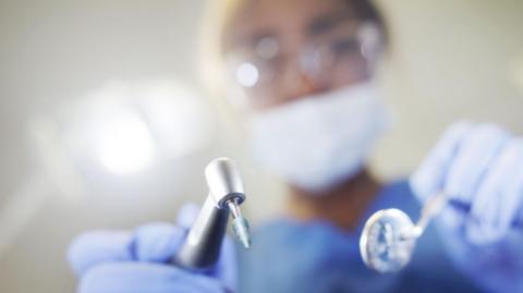 A dentist stands oversomeone with shiny metal tools. They are wearing glasses with a mask over their mouth. 