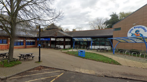 The front of a building with a sign reading "Little Kingfishers". There are blue bike rack coverings and picnic tables at the front. 