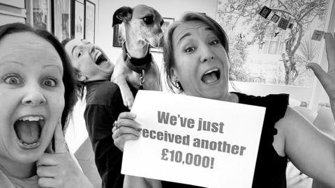 Three women grin at the camera, one of whom is holding a sign saying 'We've just received another £10,000' while another holds a dog