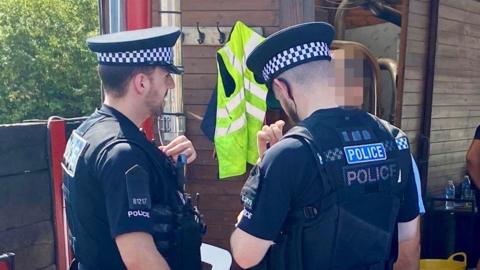 Two police officers in uniform are talking to a person whose face has been hidden from view. They are standing in front of a wooden building.