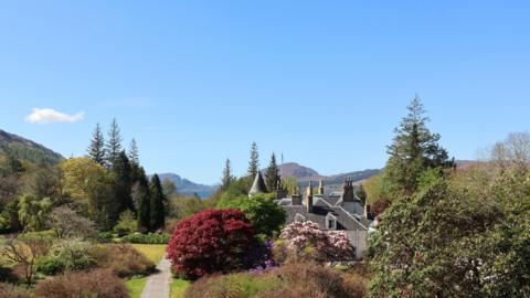 Blue sky over a large residence nestled amongst topiary and hedges with a loch in the distance
