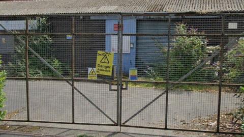 A yellow sign that reads "Danger Asbestos" is pinned to a metal gate. Behind the gate is a metal industrial unit which appears to be dilapidated with vegetation growing in front of it. 