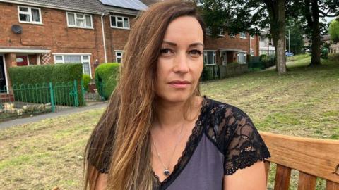A woman with long brown hair and wearing a purple and black top, sits on a wooden bench in a park, with a row of terraced houses visible behind her.