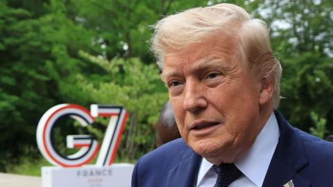 Donald Trump looking into a camera, behind him is a large sign that says G7.