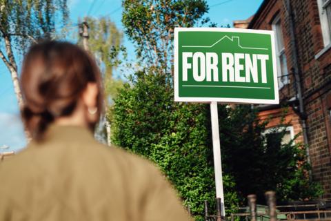 A woman blurred in the foreground looks at a green sign that says FOR RENT