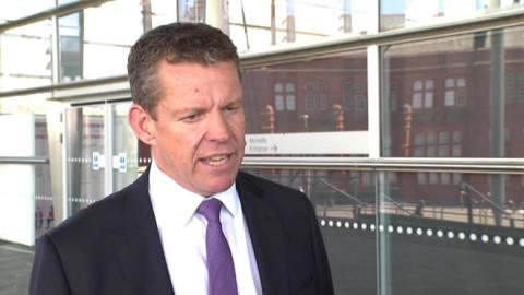 Rhun ap Iorwerth being interviewed outside the front of the Senedd building. He is wearing a dark business suit, a white shirt and a light purple tie.
