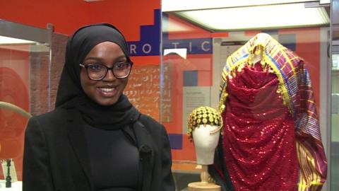 A woman wearing glasses and a black headscarf smiling in front of a glass museum exhibit.