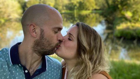 A man with a light beard wearing a light blue patterned polo shirt is to the left of the picture. A woman with shoulder length blonde hair stands beside him. They are kissing.