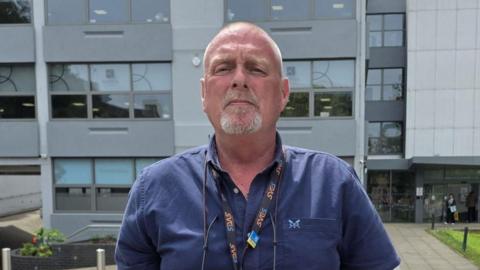 Geoff is standing outside an office building wearing a blue shirt and looking at the camera.