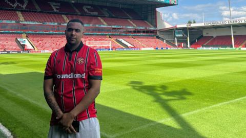 1SideJay in a Walsall FC top, standing on the pitch at the club's stadium. Behind him, there is an empty stand with red seats