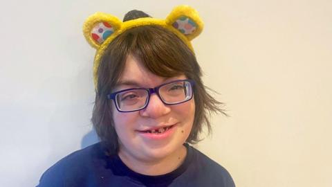 Thomas, 14, is wearing dark‑rimmed glasses and a yellow headband, and is photographed against a plain light background.