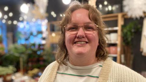 A woman with shoulder-length curly light brown hair and rectangular glasses. She is wearing a striped top and white cardigan and is smiling into the camera. The background is blurred but she is standing in a plant shop.