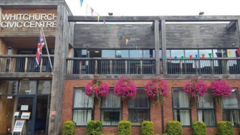 The front of a two storey building. The first floor is made of red brick and has hedges and purple hanging flowers in front of it. The second storey is mostly clad in timber. A sign on the second storey says Whitchurch Civic Centre