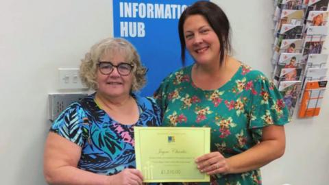 A woman with short grey curly hair stands next to a woman with dark hair. They smile at the camera and hold a yellow certificate which says 'Jayne Charles' and '£1,510,00' on it.