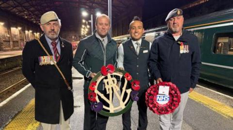 Iain Henderson from The Veterans Charity, Great Western Railway staff, and Jason Thomas holding wreaths by the Poppies to Paddington train at Penzance station. They are stood on the train platform. To the right is a GWR train. 