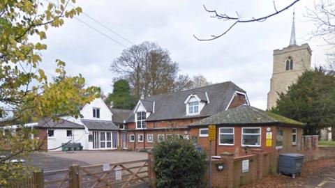 A primary school with a yard to the left and old two-storey brick buildings to the right. There is a white building with a triangular upper floor in the background, and a brick wall in the foreground with a wooden gate across the entrance. There is a church to the right.