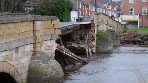 View across a swollen River Wharfe in Tadcaster showing substantial damage to the central section of a sandstone bridge. In the background there are Georgian buildings lining the town's high street.