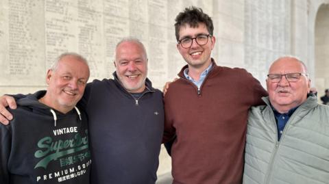 Four men are smiling at the camera. They are standing in front of a wall with names inscribed. They are all smiling and have their arms around each other. 