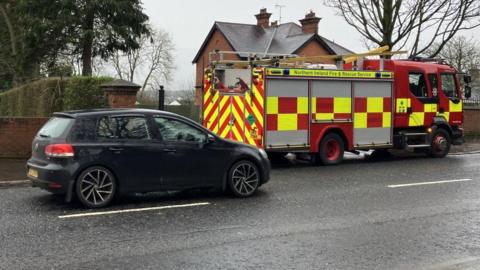 Shows a fire appliance in red and yellow in front of a house. A black car is to the left of the photograph.