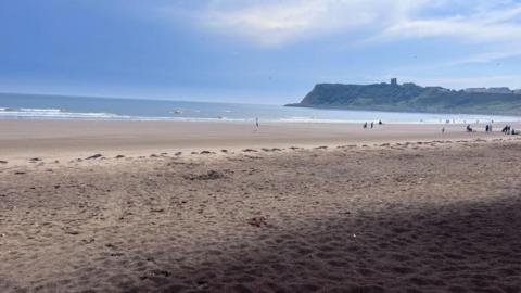 A view of North Bay in Scarborough, a stretch of sandy beach with the sea in the background. In the distance, a ruined castle can be seen on cliff tops.