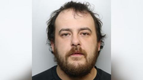 A police mugshot of a man with messy dark hair and a short beard. He is wearing a black T shirt.