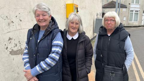 Three middle-aged women are standing together in front of a dirty cracked white wall. They all have grey hair and are wearing black jackets with blue tops under them. 