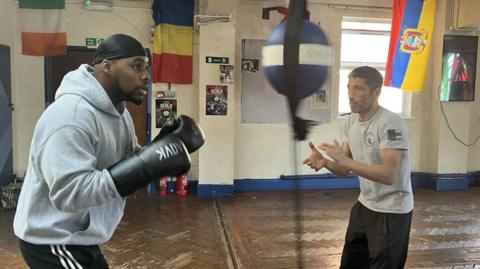 A man in a boxing gym wearing black boxing gloves and a grey hoodie takes aim at a punching bag as another man to the side claps in encouragement. 