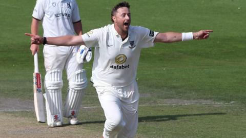 Ollie Robinson runs down the wicket celebrating with both arms aloft, wearing Sussex's all white kit, with a batter standing in the background