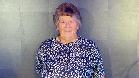 Jo Norman is wearing a navy blue and white patterned short-sleeved top.
She is smiling at the camera and has short brown hair.