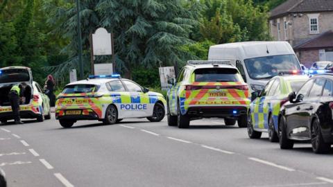 Four police cars can be seen in the middle of a road, with an officer looking in the boot of one of them. A white van can be seen parked up next to it with trees and housing in the background.