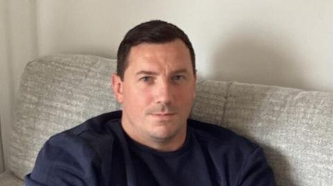 A man sitting on a grey sofa, wearing a blue jumper and a serious expression 