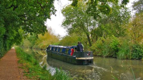 A narrowboat on green river water surrounded by green trees and bushes, with a river path alongside