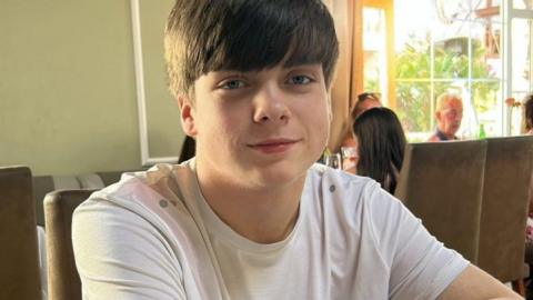 Harry Begg sits at a table in a restaurant, he has short brown hair with a fringe. He wears a white t-shirt