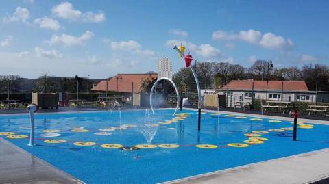 The splash park in Douglas. It has a blue ground, water sprays up, and there are arches to run through, there are buildings in the background.