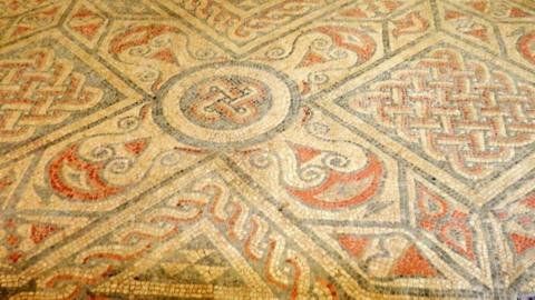 A close up of the mosaic floor which uses blue-grey, red, yellow and cream colours. The pattern has a knot-like design.