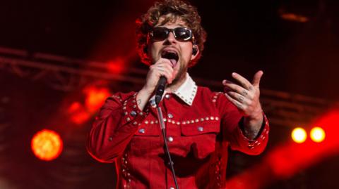 Tom Grennan, on the stage wearing a red jumpsuit, with one arm out and the other holding a microphone. He has short curly hair and is wearing sunglasses. Stage lights are behind him.