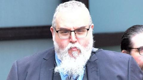 A man with glasses, short grey hair and a long white beard looks at the camera. He is wearing a three-piece grey suit and tie, with a blue shirt. 