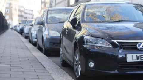 A close up image of a row of dark-coloured cars parked on a street next to a slab pavement.