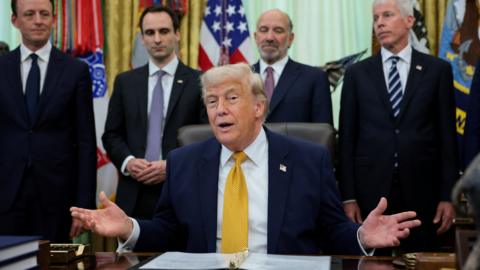 A man in a suit sits at a desk with four men standing behind him, his hand held open. 