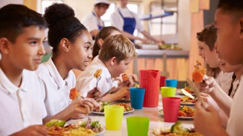 Several primary school pupils sit at a canteen table eating a meal. They are all wearing white uniform polo shirts and are smiling at one another. Alongside the food in front of them are multi-coloured drinking cups and a jug.