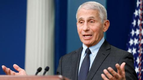 Anthony Fauci speaking at a podium in front of a US flag wearing a grey suit and blue tie with dots.