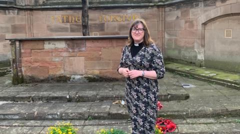 A female reverend in a long black floral dress and black rimmed glasses standing in the ruins of a cathedral, smiling at the camera