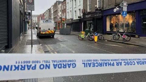 Garda tape in the forefront of the picture in a street in Dublin. A police van is further back in the street as are three parked bicycles and shops.