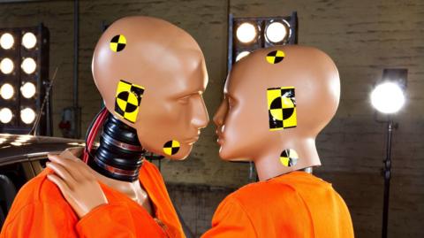 Two crash test dummies in an embrace with lights behind them