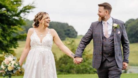 A newlywed couple hold hands and smile at each other against a backdrop of hills and trees. The groom is wearing a grey suit and the bride, who is holding some flowers in her other hand, is wearing a traditional white dress.