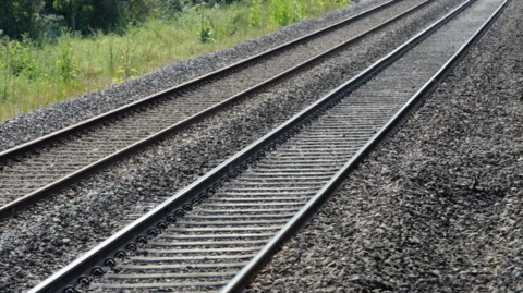 Two railway lines can be seen with gravel underneath and either side of them. Grassland is visible on the left side of the tracks.